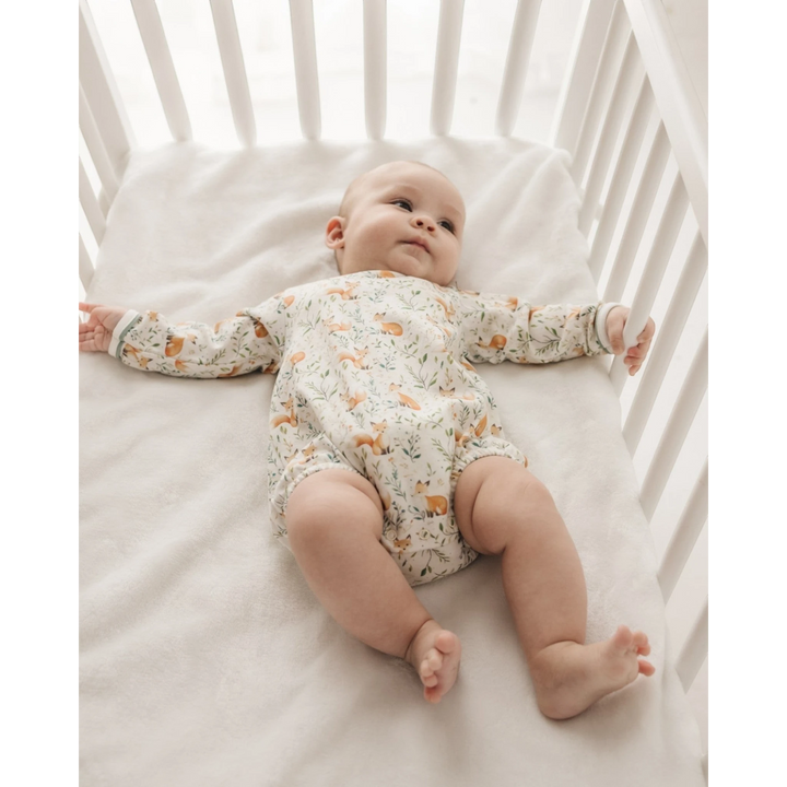 A baby lying on their back in a white wooden crib, wearing a long-sleeve cream-colored romper with a delicate orange and green animal print, that store