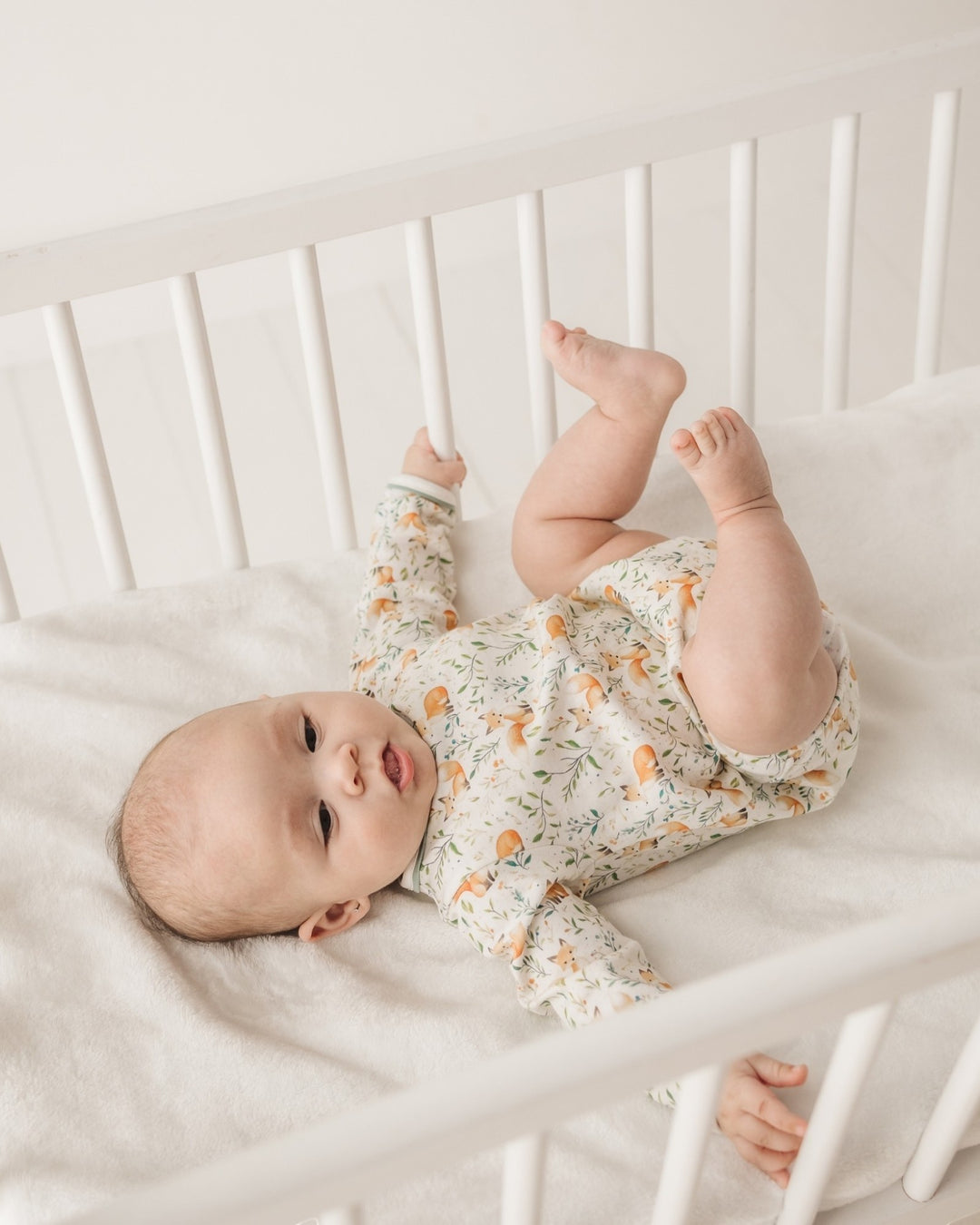 A baby lying on their back in a white wooden crib, wearing a long-sleeve cream-colored romper with a delicate green and orange floral pattern, that store