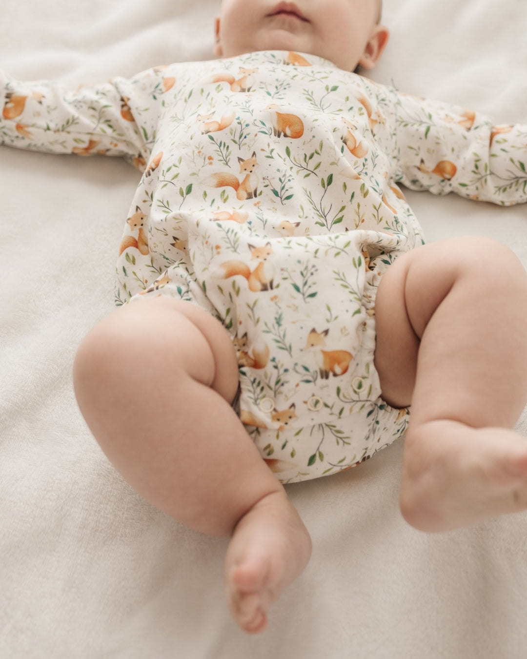 A baby lying on a soft cream-colored blanket, wearing a long-sleeve cream romper printed with small orange foxes and green leaves, that store