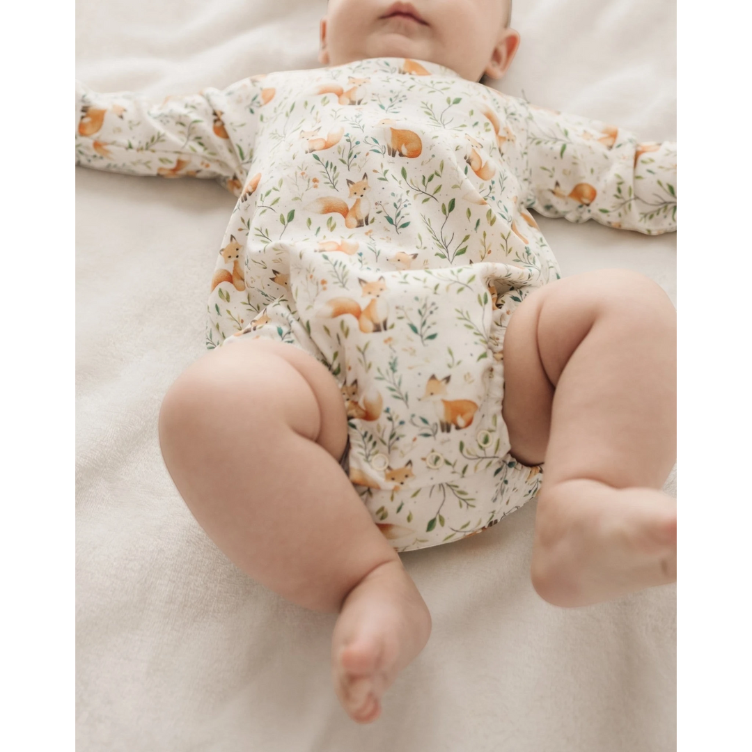 A baby lying on a soft cream-colored blanket, wearing a long-sleeve cream romper printed with small orange foxes and green leaves, that store