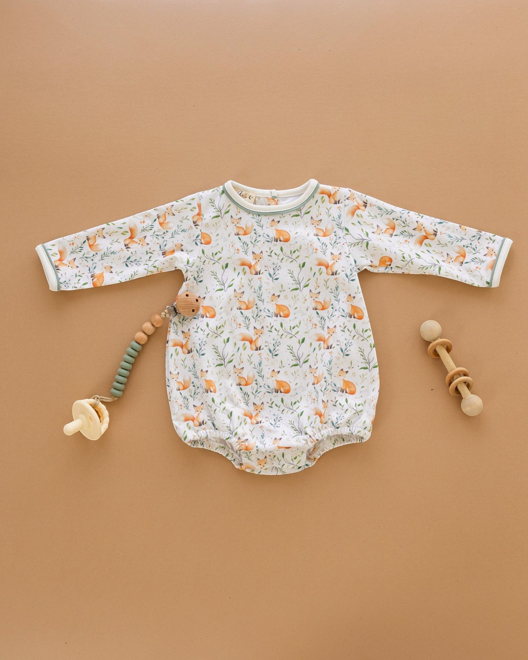 A baby long-sleeve bodysuit with an all-over fox and leaf print, laid flat on a warm tan background with baby toys nearby.
