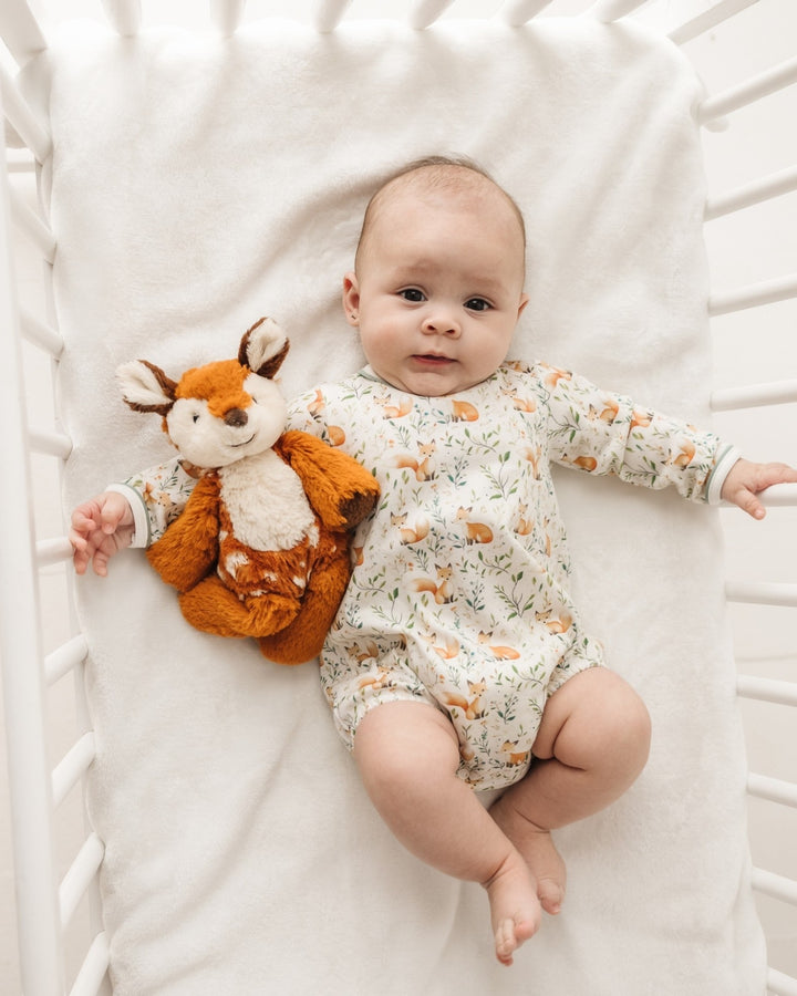 A baby lying on a white blanket inside a white crib, wearing a long-sleeve cream romper with a small orange and green woodland animal print, and holding a plush orange fox toy, that store
