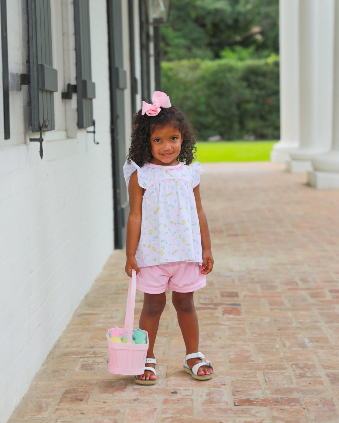 Young girl in a white blouse and pink shorts holding a pink basket with Easter eggs on a brick path.