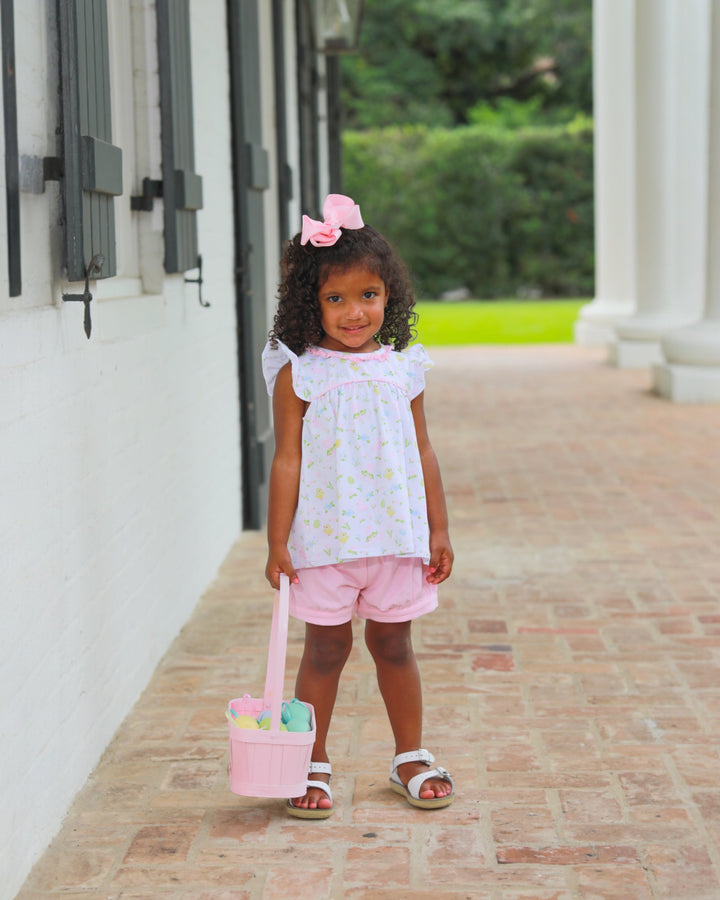 Young girl in a white blouse and pink shorts holding a pink basket with Easter eggs on a brick path.