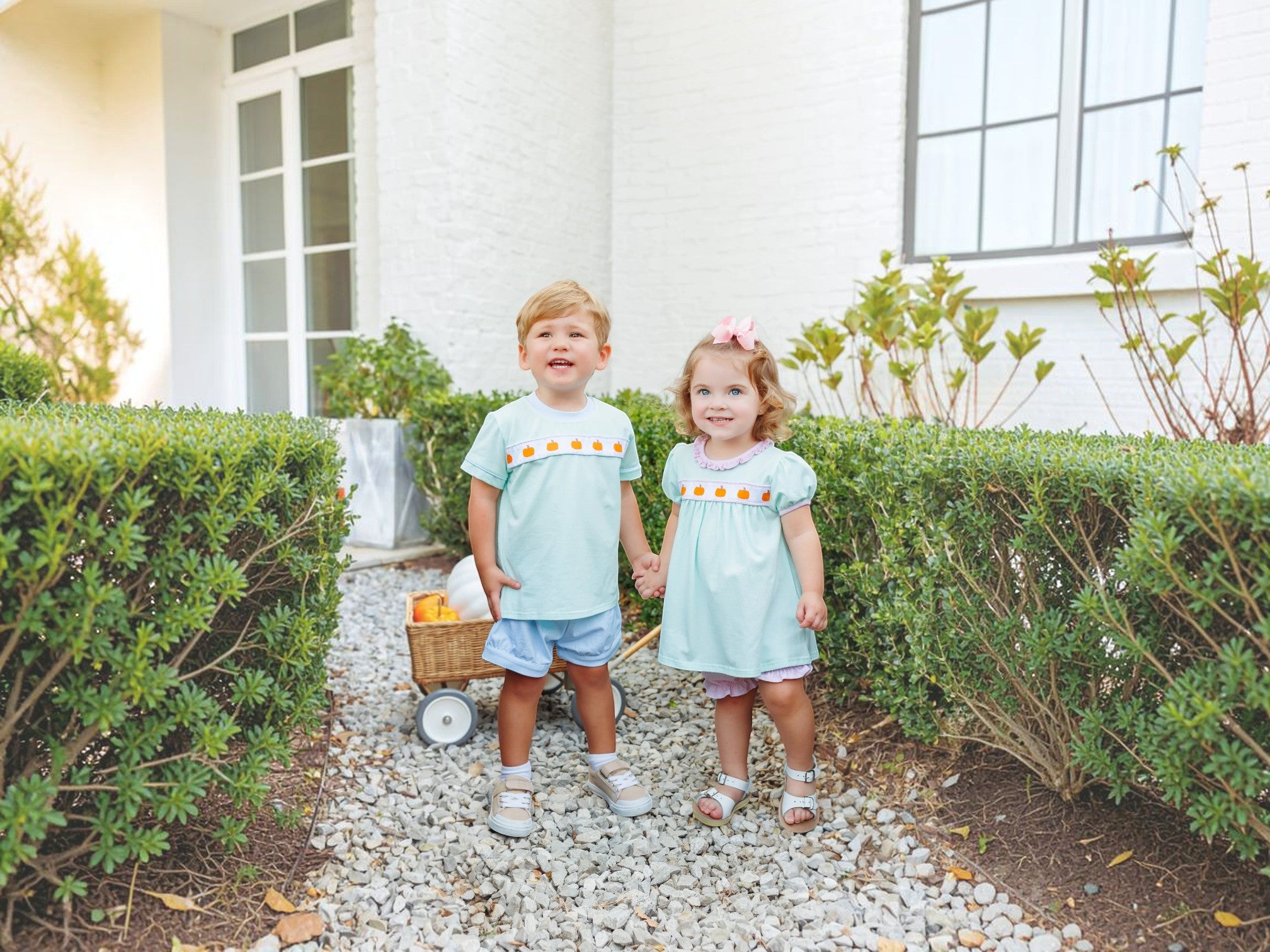 Two young children standing on a gravel path between green hedges in front of a white house. The child on the left wears a light blue smocked shirt and denim shorts, and the child on the right wears a matching light blue smocked dress with a pink bow, that store
