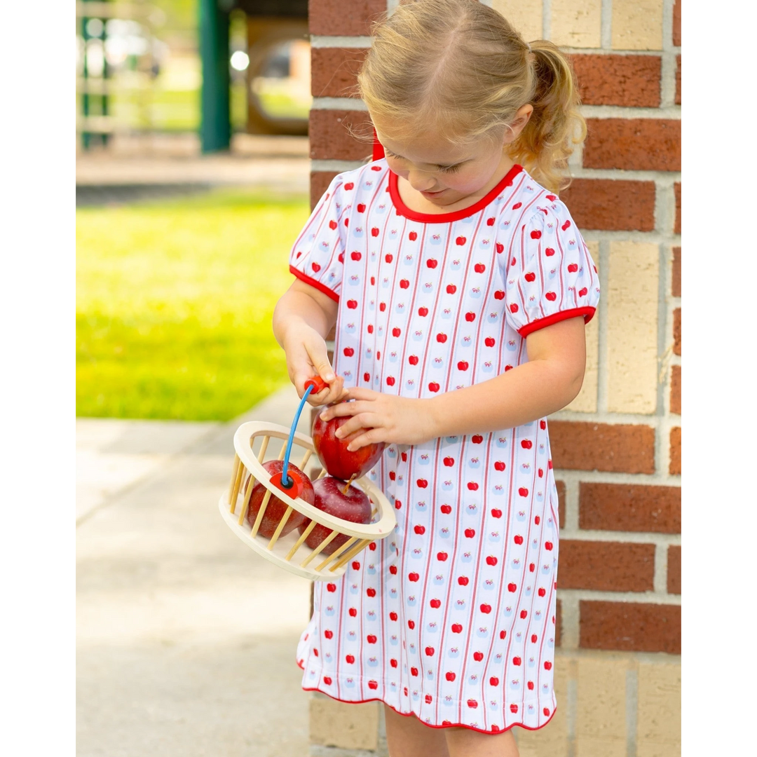 A young girl wearing a That Store apple print dress with red trim, holding a small basket of apples outdoors.