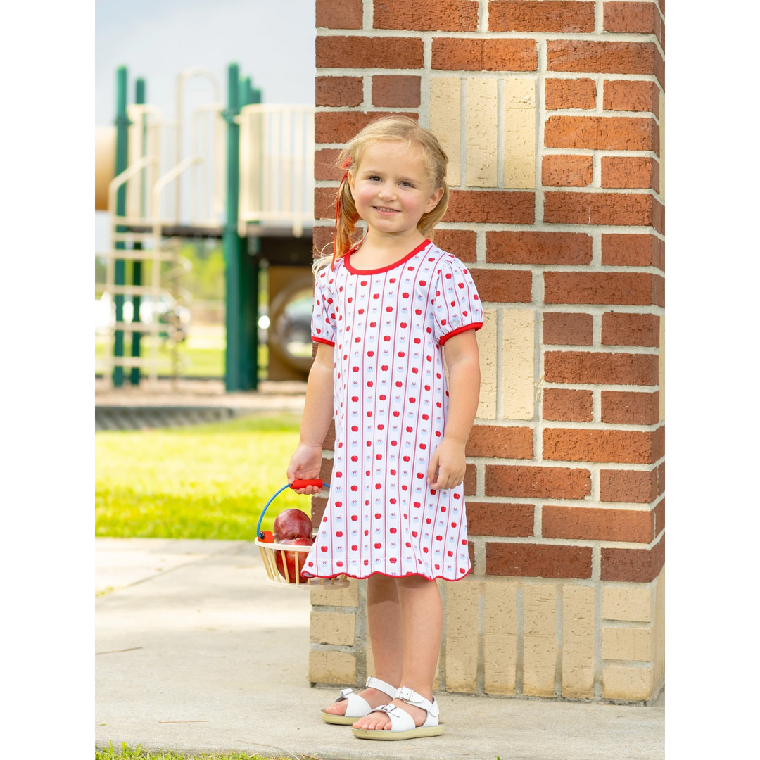 A young girl wearing a That Store apple print cotton dress with red trim, standing near a brick wall at a playground.
