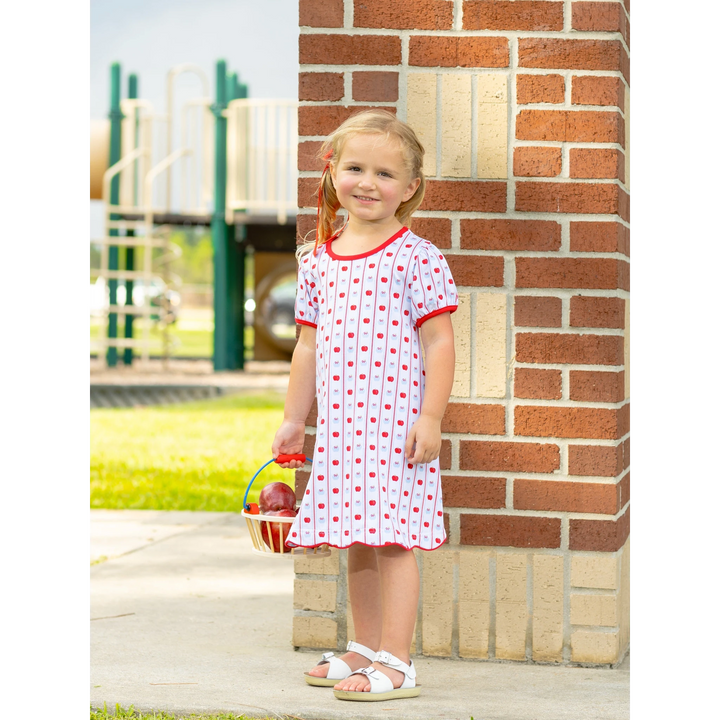 A young girl wearing a That Store apple print cotton dress with red trim, standing near a brick wall at a playground.