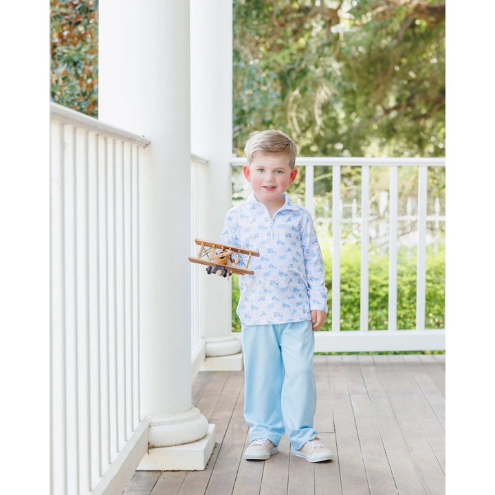 Young boy wearing a That Store blue airplane print 1/4 zip pullover and light blue pants on a porch holding a toy plane