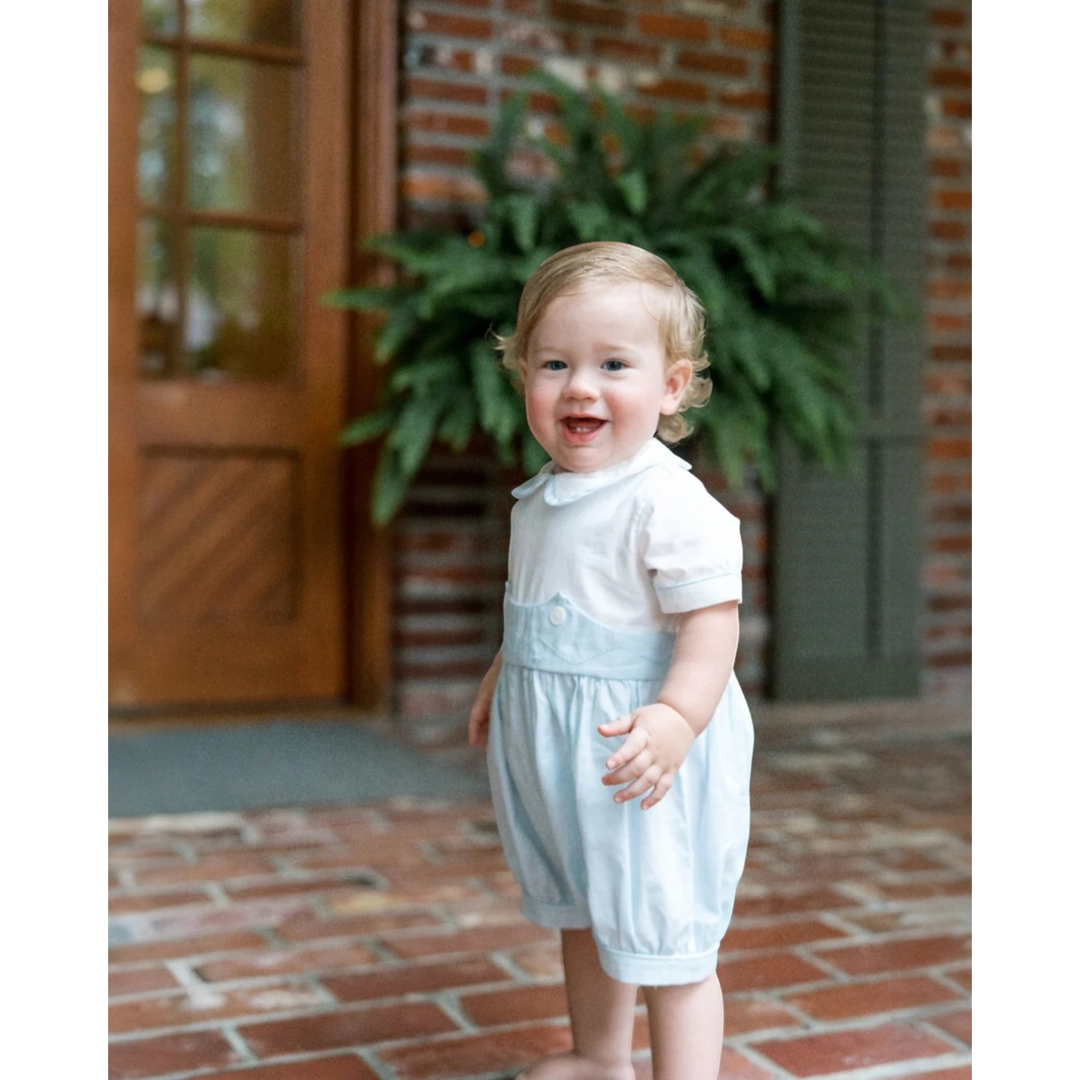 That Store baby boy wearing a light blue and white batiste bubble romper for special occasions on a brick porch.