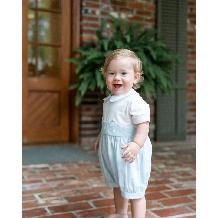 That Store baby boy wearing a light blue and white batiste bubble romper for special occasions on a brick porch.
