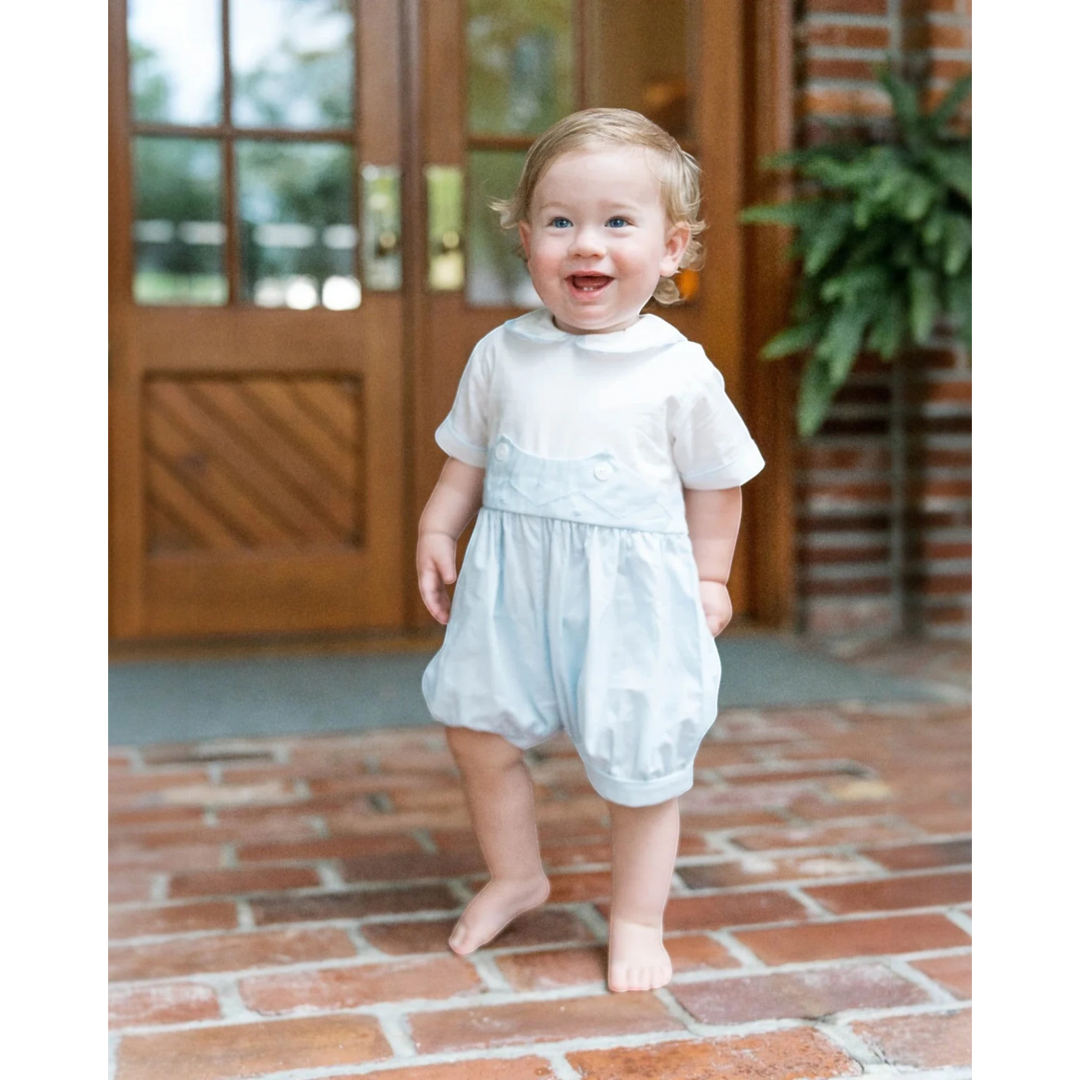 Toddler boy wearing a That Store light blue batiste bubble romper with a white collar standing on a brick patio.