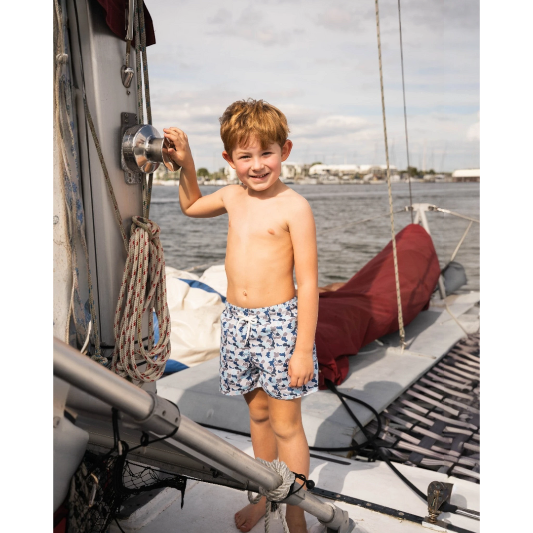 Young boy wearing That Store blue camo Saint Simons swim trunks while standing on a sailboat deck.
