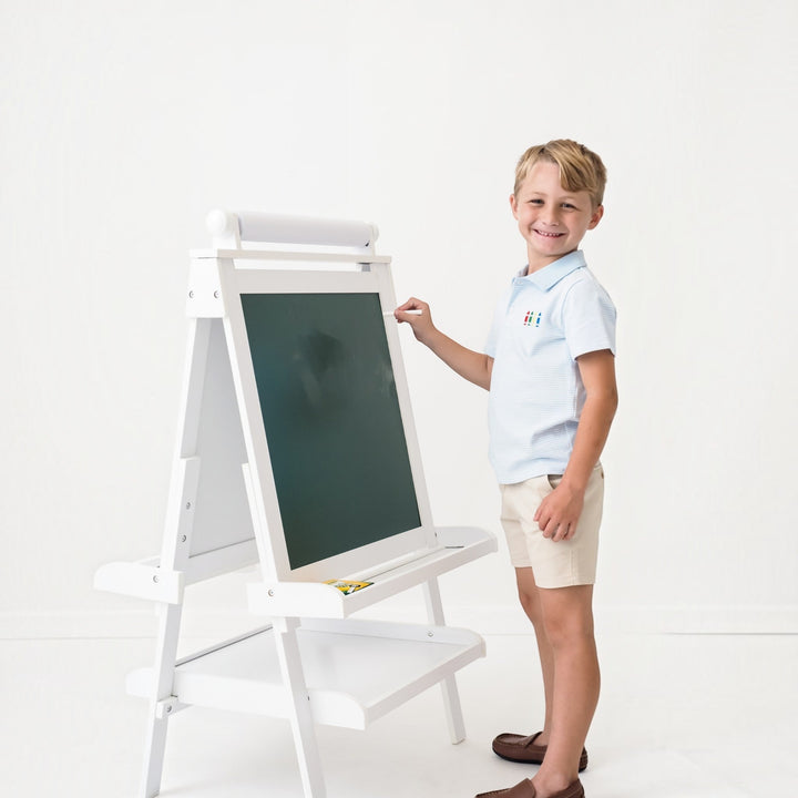 That Store boys blue stripe polo shirt with crayon embroidery worn by a smiling boy drawing on a white easel