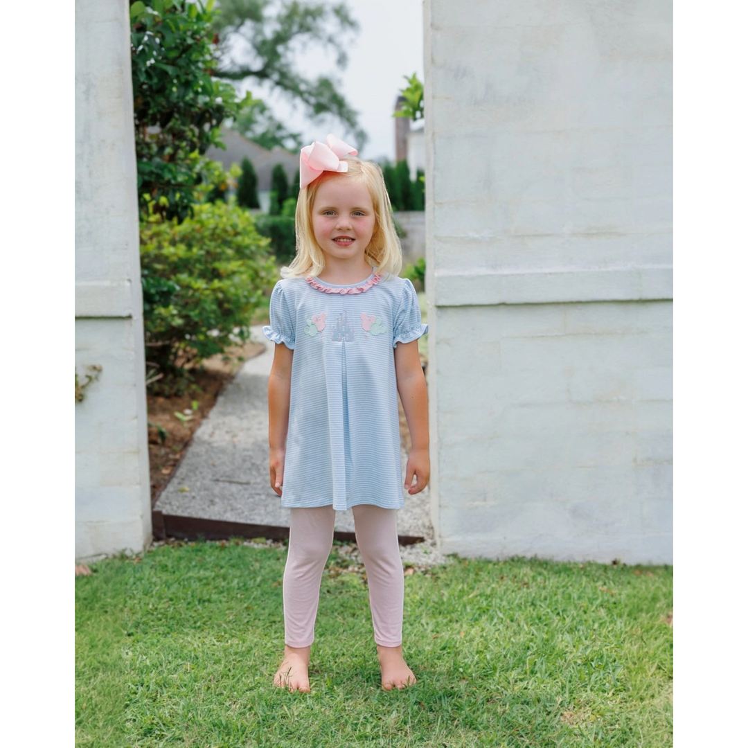 A young girl wearing a That Store blue striped tunic with castle embroidery and pink ruffle leggings outdoors.