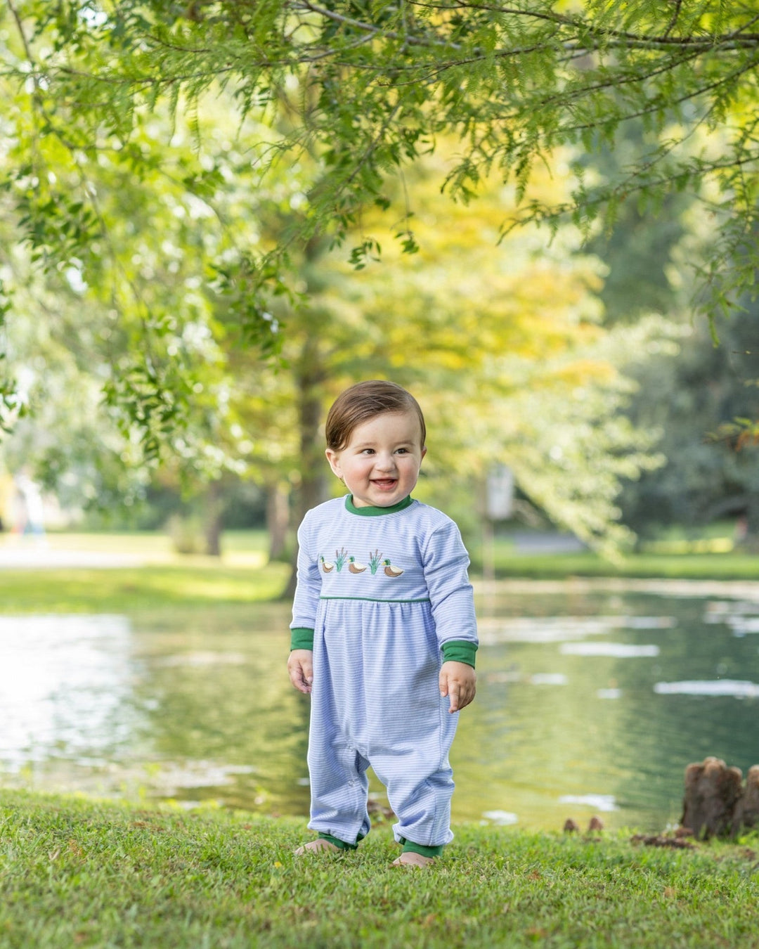 A toddler wearing a That Store blue striped cotton romper with mallard duck embroidery, standing in a park by a pond.