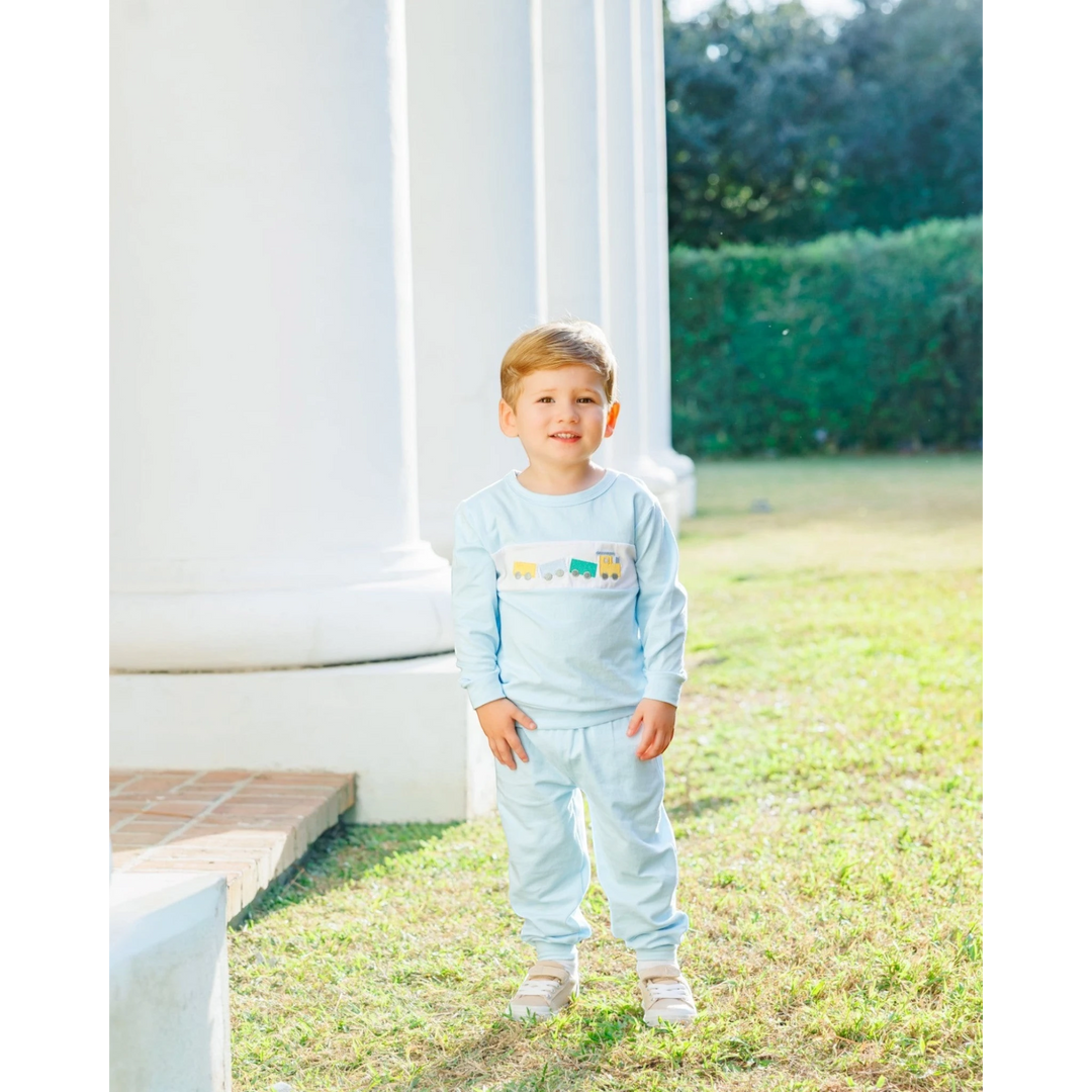 A young boy wearing a That Store light blue jogger set with a whimsical embroidered train design on the chest.