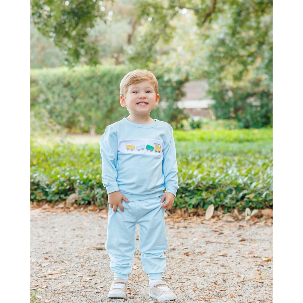 A young boy wearing a That Store light blue jogger set with a whimsical embroidered train design on the chest.