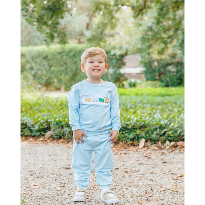 A young boy wearing a That Store light blue jogger set with a whimsical embroidered train design on the chest.