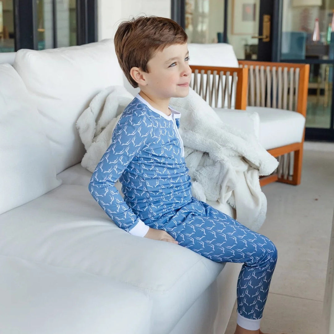 A young boy sitting on a white sofa wearing a That Store blue pajama set with a white rustic antler print pattern.