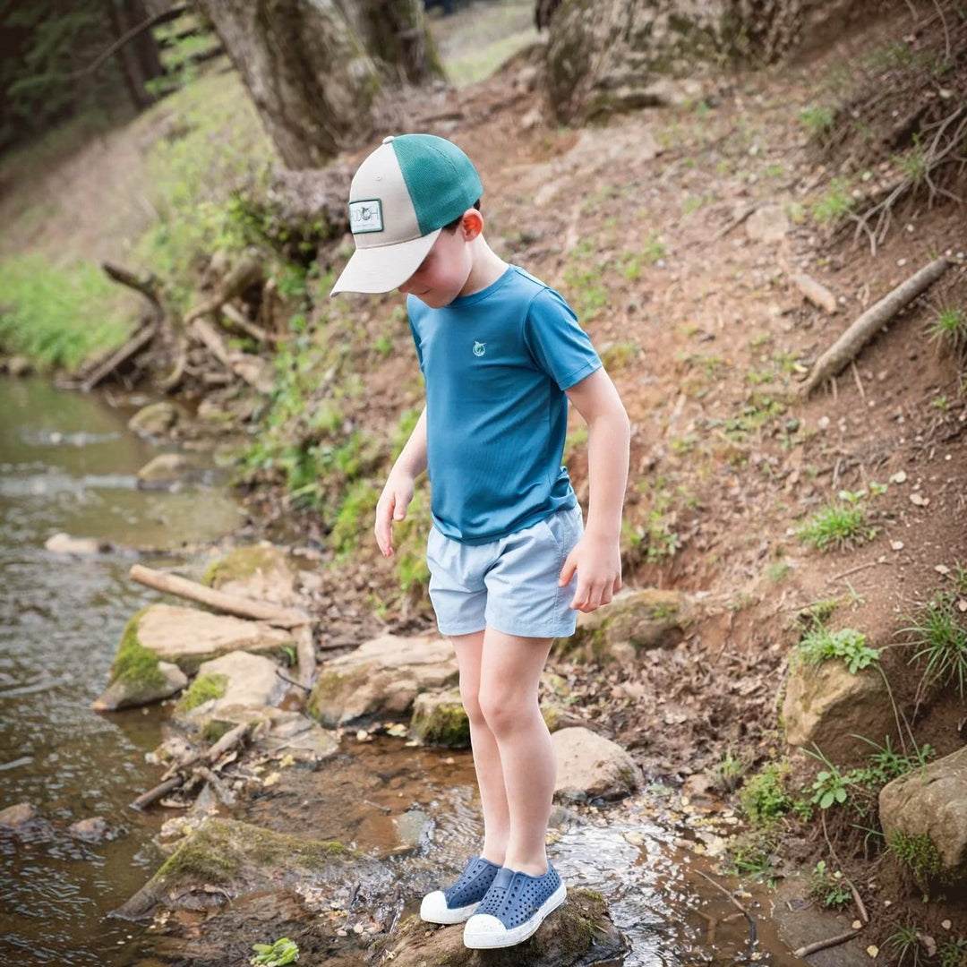 That Store boy wearing a blue performance t-shirt with UPF 50+ protection, light shorts, and a cap by a creek.
