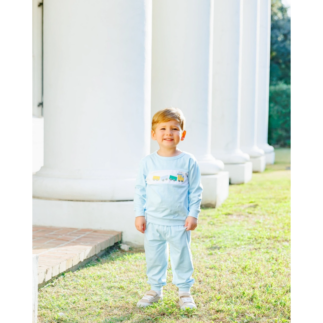 That Store boys blue jogger set with embroidered train motif worn by a young boy standing outdoors by white columns.