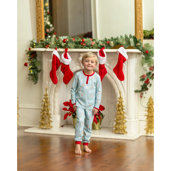 A young boy wearing a That Store blue and red cotton holiday pajama set in front of a decorated Christmas fireplace.