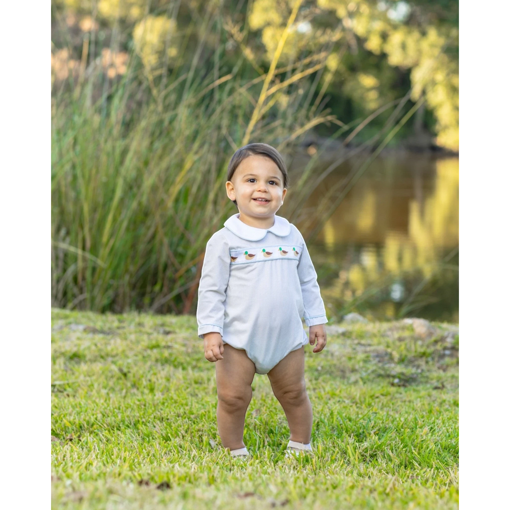 That Store boys bubble in light brown gingham with a mallard duck ribbon detail, worn by a toddler in a park.