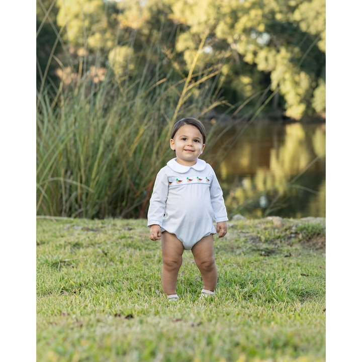 That Store boys bubble in light blue with a mallard duck ribbon trim, worn by a toddler in a park setting.