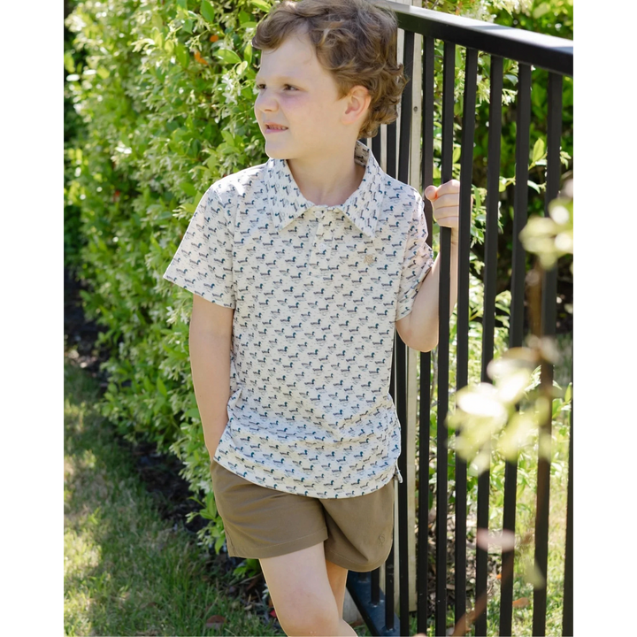 That Store boys performance polo in cream with mallard duck print, worn by a boy standing by a black metal fence.