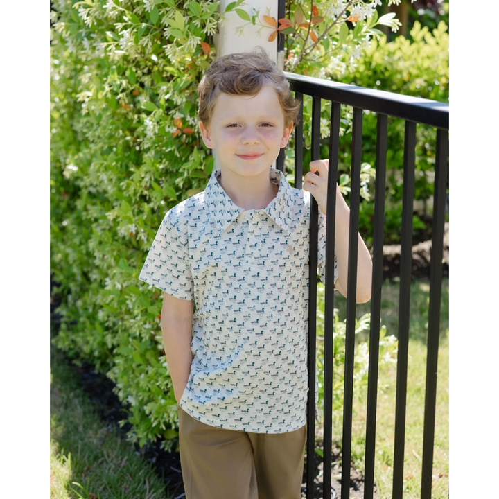 A young boy wearing a That Store cream performance polo with a mallard duck print, standing by a black metal fence.