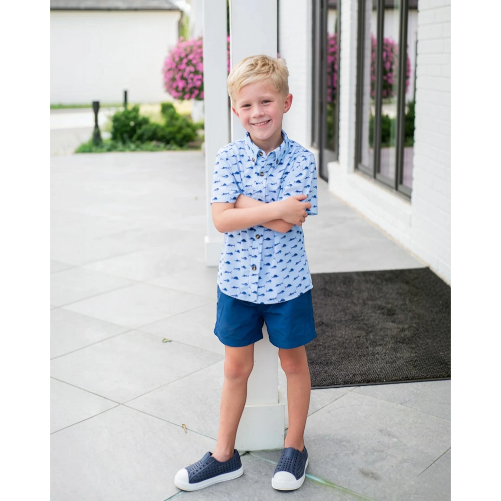 Young boy wearing That Store navy performance shorts and a patterned button-down shirt while standing outdoors.