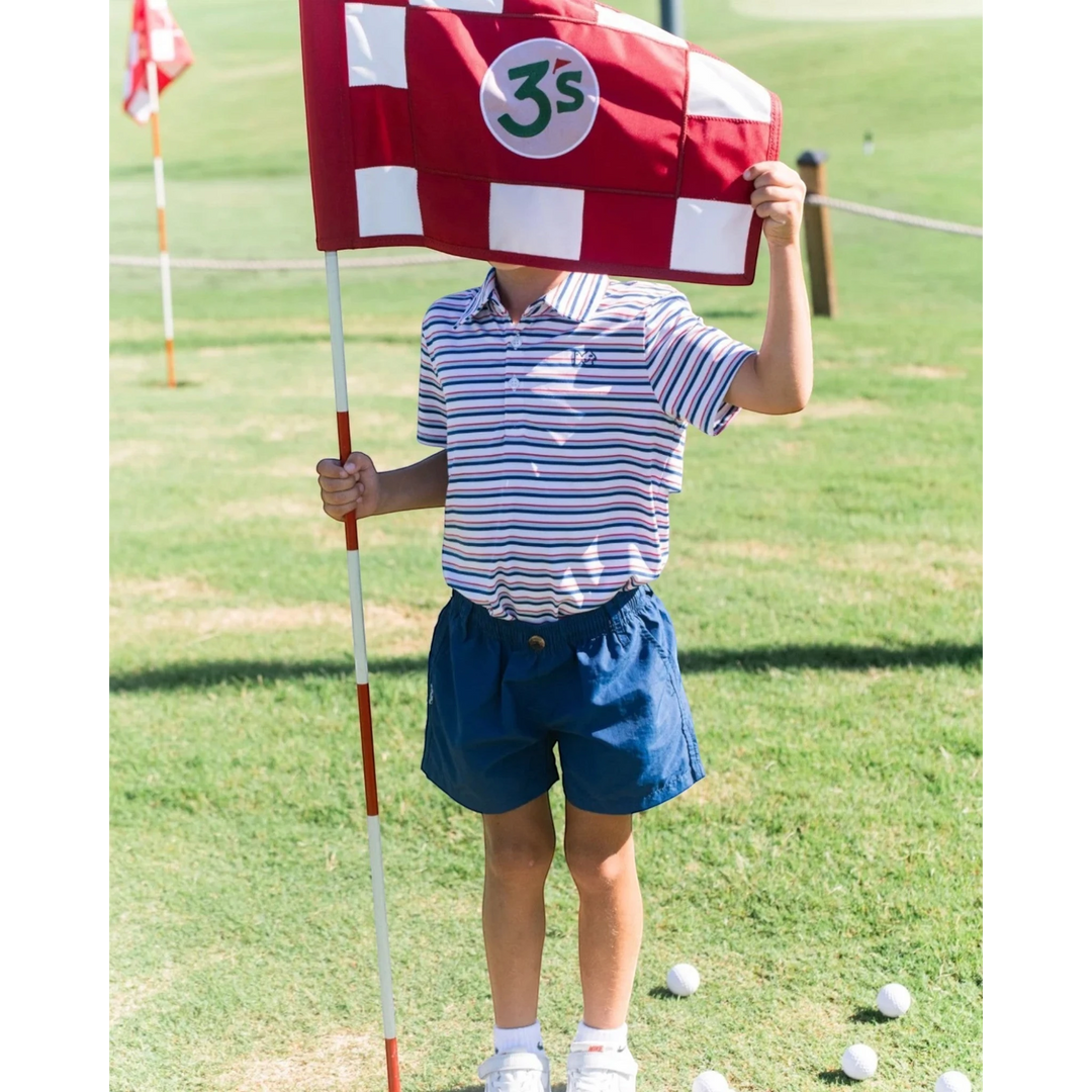 A young boy wearing That Store navy performance shorts and a striped polo shirt holding a red and white golf flag