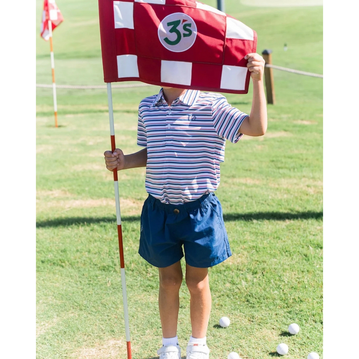 A young boy wearing That Store navy performance shorts and a striped polo shirt holding a red and white golf flag