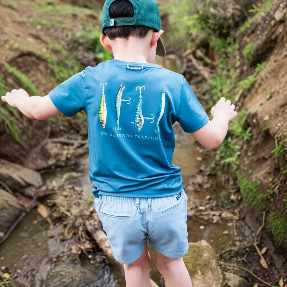 That Store boys' blue performance t-shirt with fishing lure graphic on back, worn by a child in a nature setting.