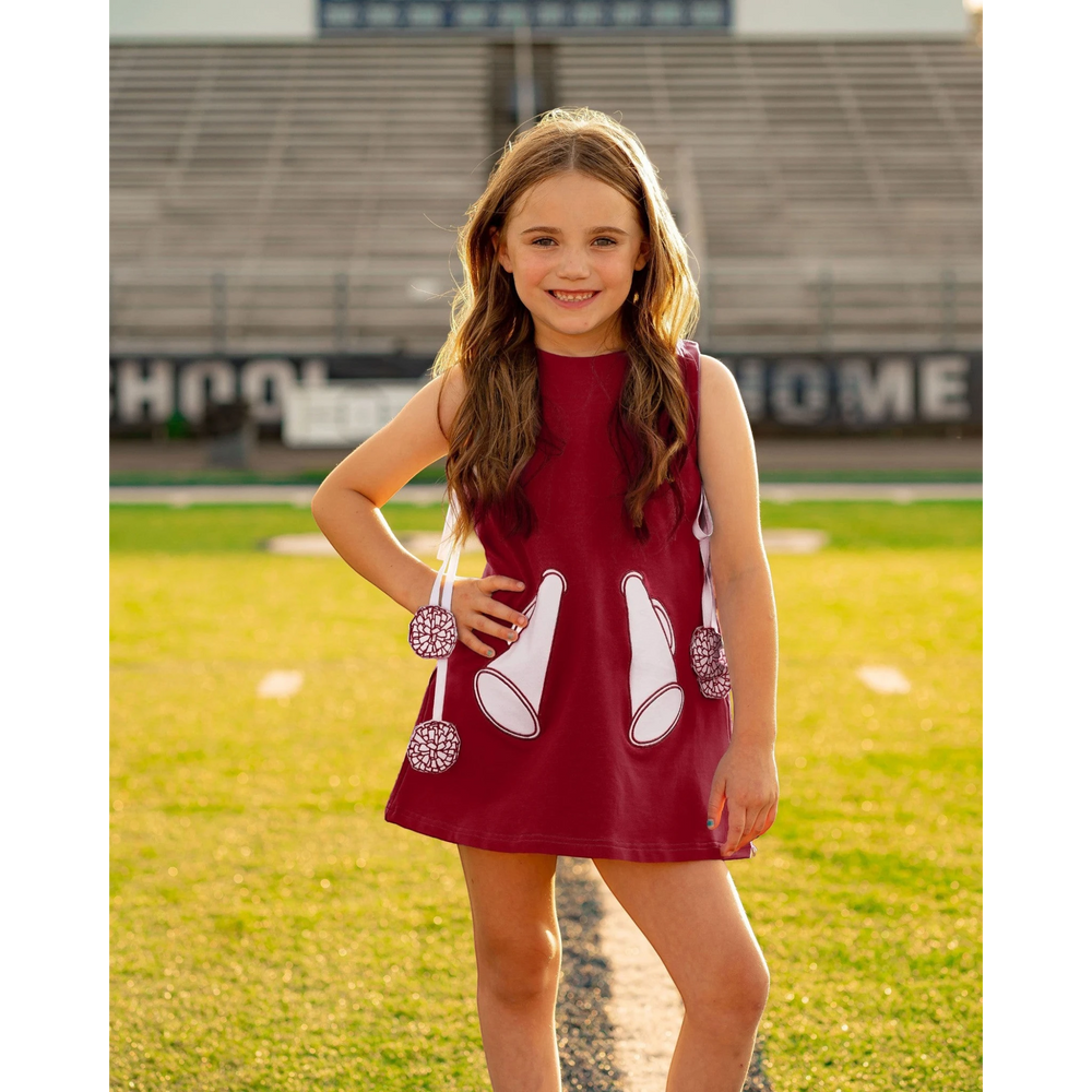 That Store burgundy cotton jumper dress with megaphone appliques and pom-pom ties worn by a girl on a football field.