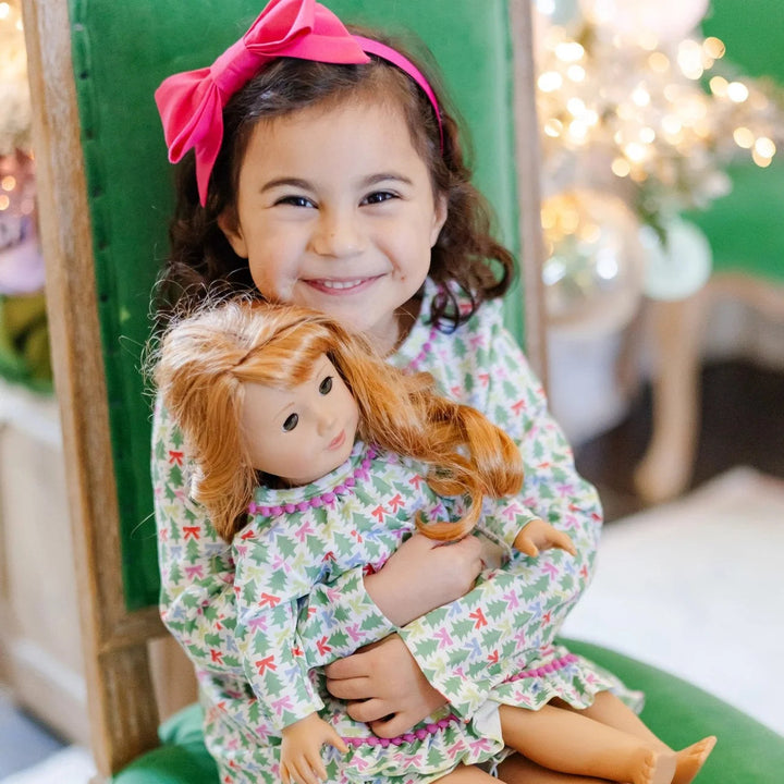 A smiling girl holding an 18-inch doll wearing a That Store Carlin dress with green trees and colorful pom-pom trim.