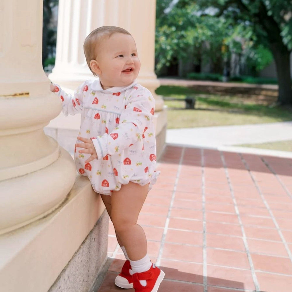 That Store Covington bubble in Barnyard Tales print with light blue piping worn by a baby standing by a white pillar.