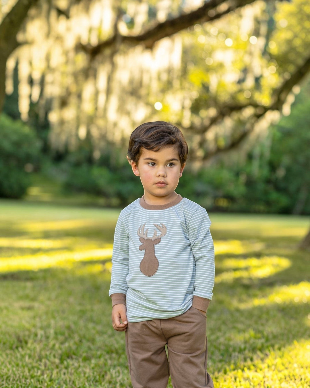 Toddler boy wearing a That Store green striped long-sleeve shirt with a deer applique and brown jogger pants outdoors.