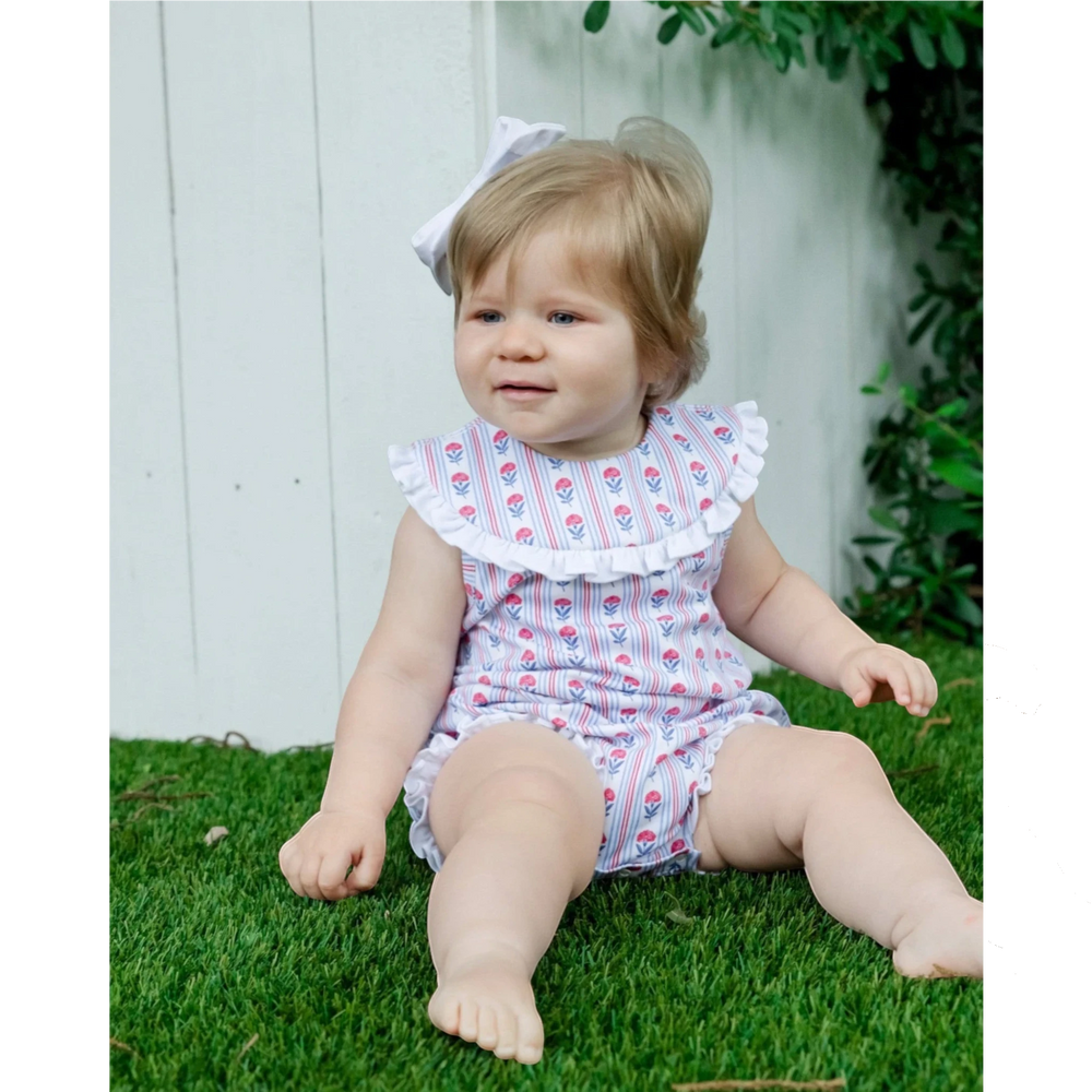 Baby girl wearing a That Store red, white, and blue floral cotton bubble with a ruffled collar, sitting on grass.