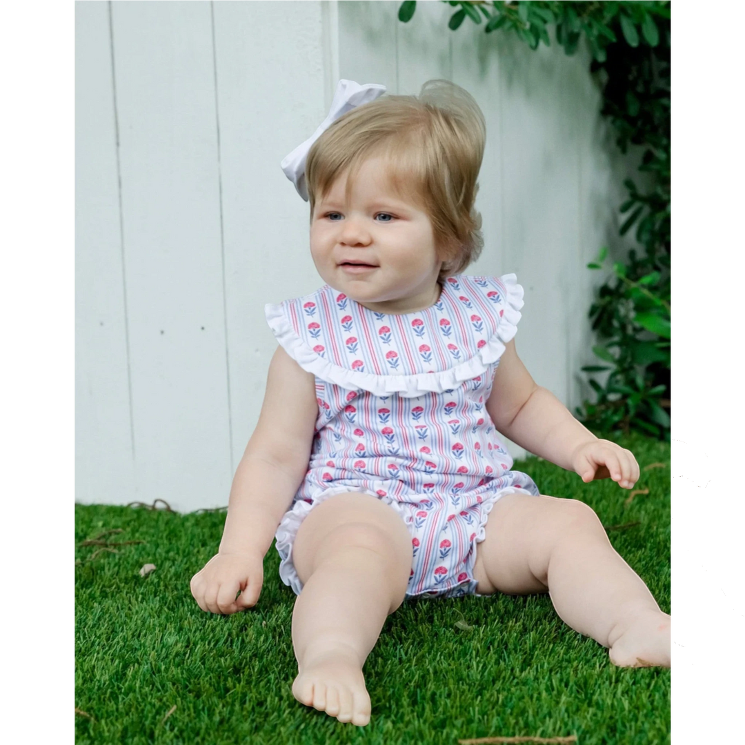 Baby girl wearing a That Store red, white, and blue floral cotton bubble with a ruffled collar, sitting on grass.
