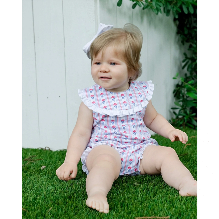 Baby girl wearing a That Store red, white, and blue floral cotton bubble with a ruffled collar, sitting on grass.