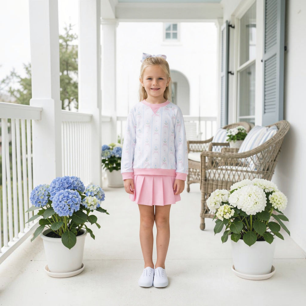 Young girl wearing a That Store pink and white cameo print pullover and matching pleated skirt on a porch.