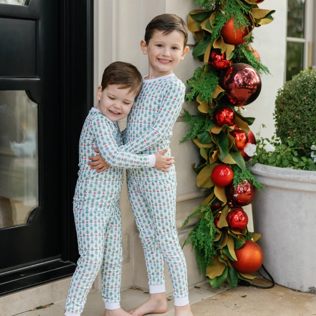 Two boys wearing That Store Grayson pajama sets with green trees and red stars, standing by festive holiday decor.