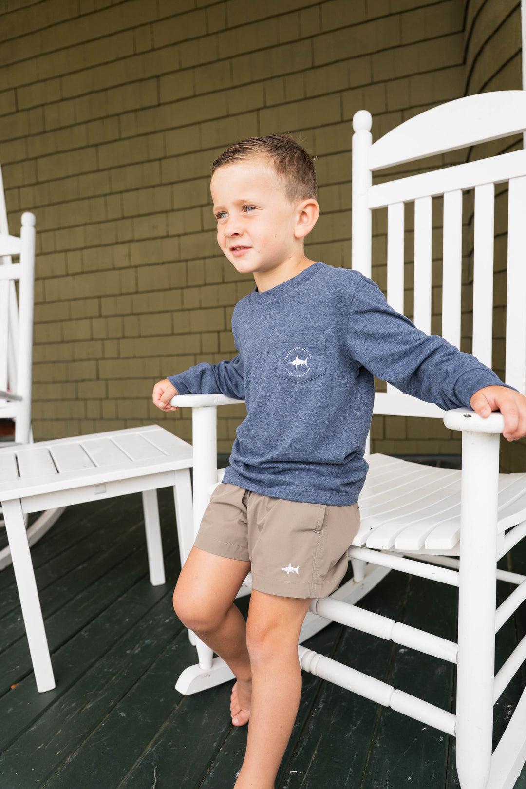 Young boy wearing a That Store heather blue long sleeve tee with a small chest graphic, paired with tan shorts.