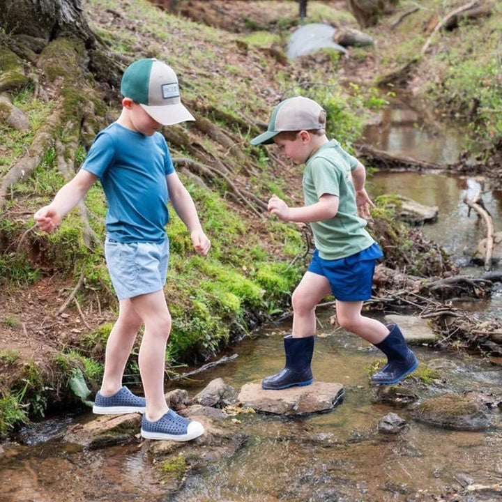 Two boys wearing That Store hedge green cotton t-shirts and baseball caps playing in a shallow forest stream.
