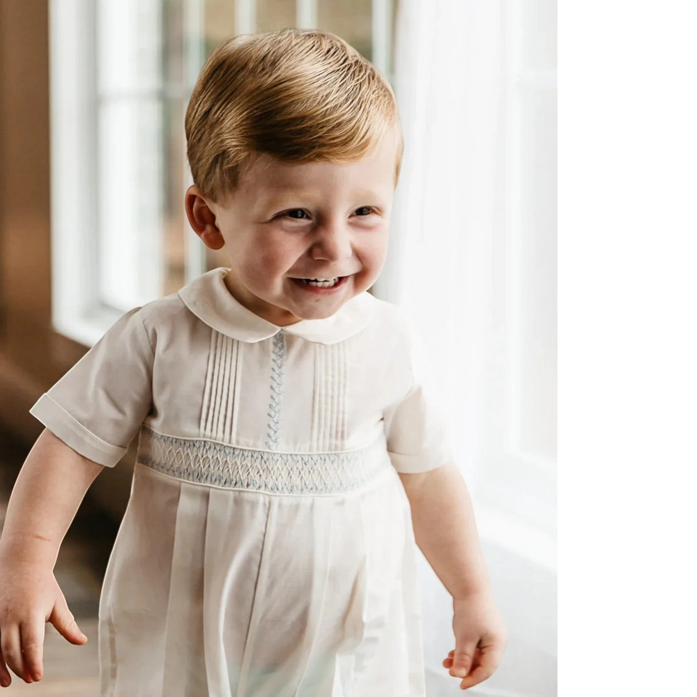 That Store ivory boys romper with hand-stitched pintucks, blue embroidery, and French lace collar on a smiling toddler