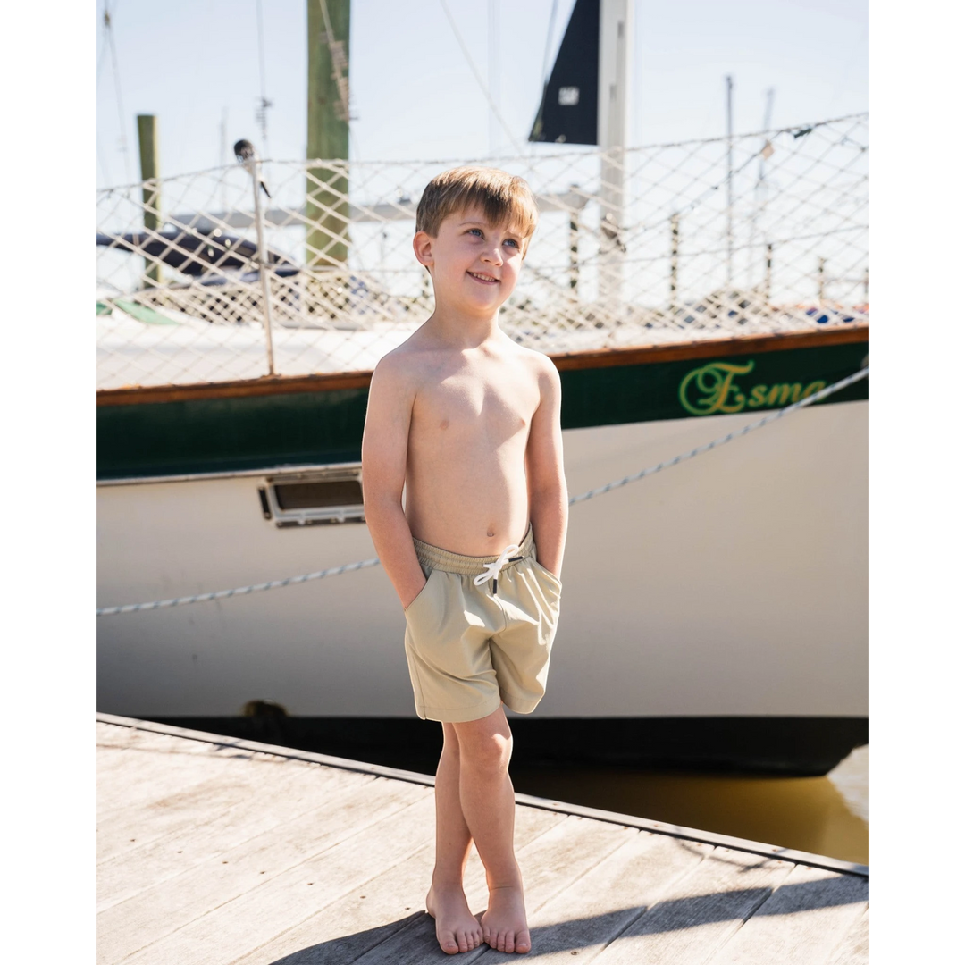 Young boy wearing That Store khaki performance shorts with an elastic drawstring waist on a wooden dock by a sailboat.