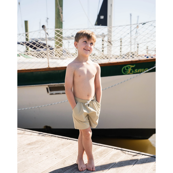 Young boy wearing That Store khaki performance shorts with an elastic drawstring waist on a wooden dock by a sailboat.