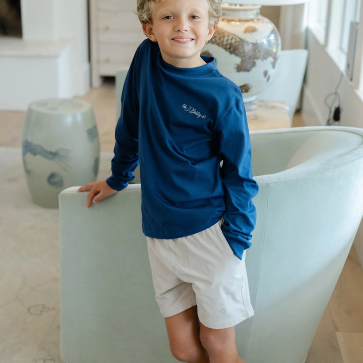 Young boy wearing That Store khaki performance shorts and a blue long-sleeve shirt, standing in a bright room.